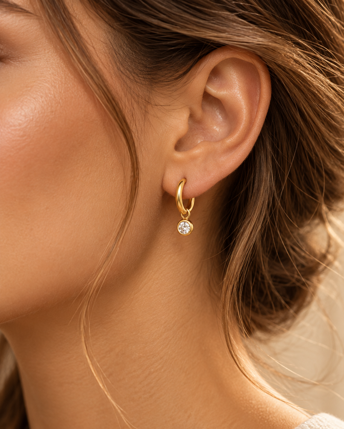 Gold tone stainless steel huggie earrings with a bezel-set clear stone drop, close up